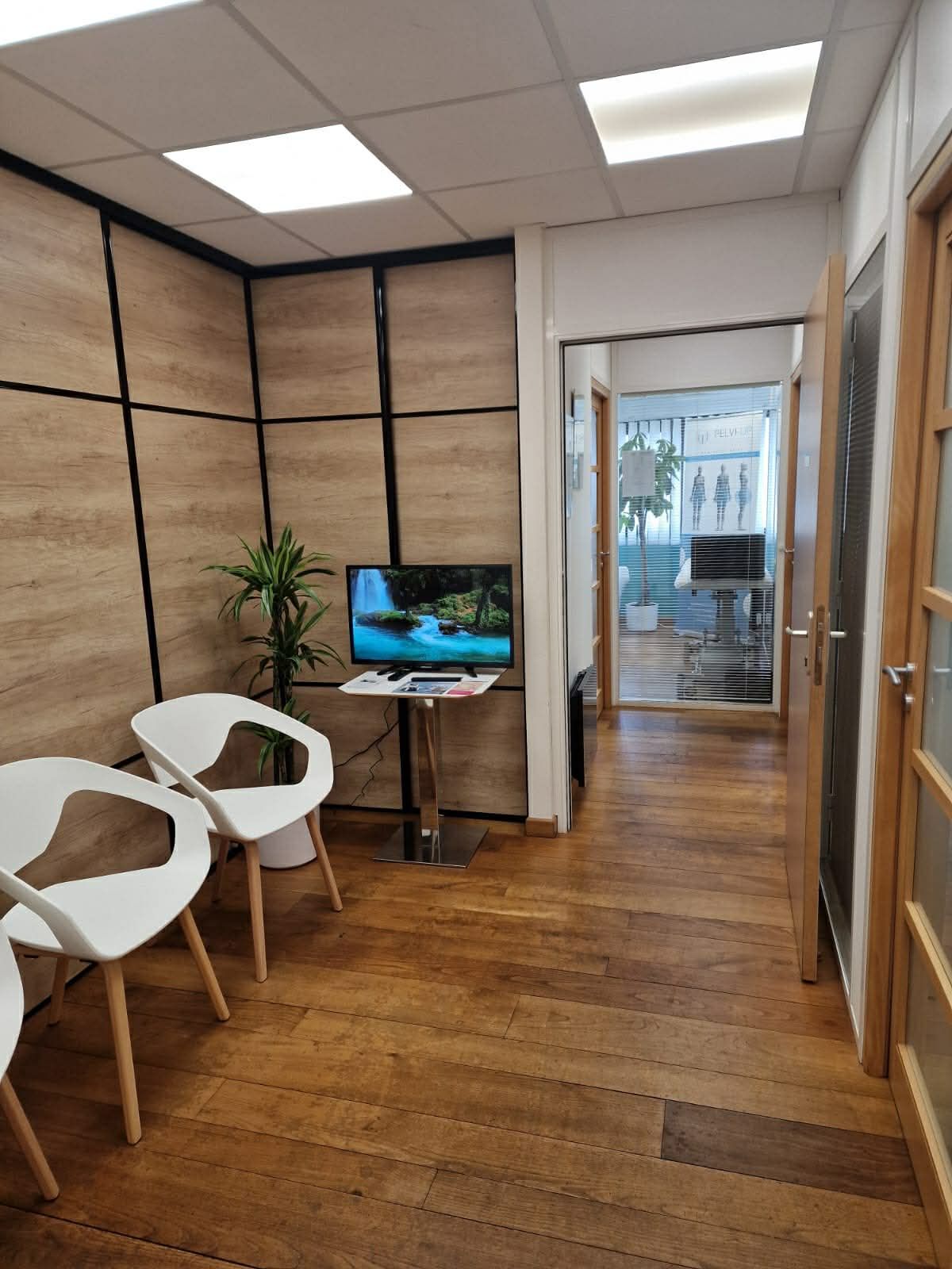 Une salle d'attente avec deux chaises blanches, un petit écran et une plante en pot, donnant sur un espace de bureau.
