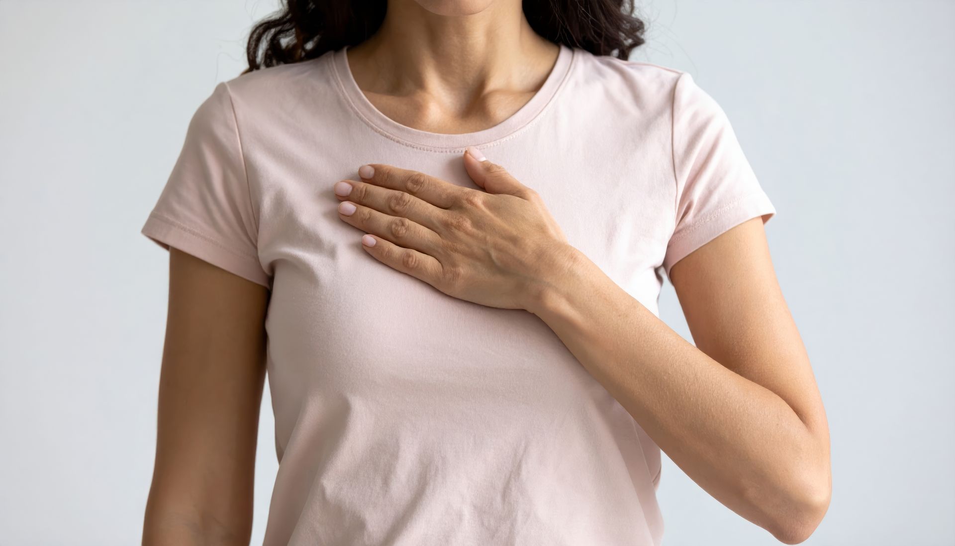 Une personne vêtue d'un t-shirt rose clair pose sa main droite sur sa poitrine.