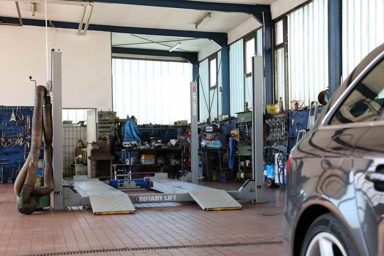 Ein Auto steht auf einer Hebebühne in der Werkstatt eines Mechanikers; Werkzeuge hängen an der Wand, helle Beleuchtung.