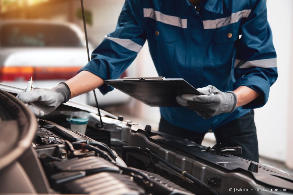 Ein Mechaniker in blauer Arbeitskleidung inspiziert mit einem Klemmbrett in der Hand und Handschuhen den Motor eines Autos.