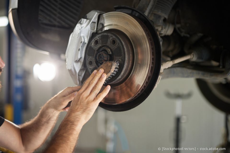 Ein Mechaniker inspiziert die Bremsscheibe eines Autos. Seine Hände berühren die Bremsscheibe, der Bremssattel ist sichtbar. Die Szene spielt in einer Werkstatt.