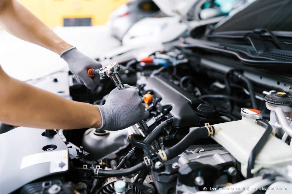 Hände mit grauen Handschuhen arbeiten mit einem Schraubenschlüssel an einem Automotor.