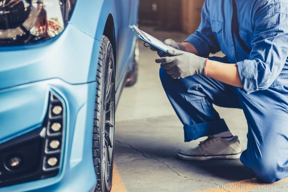 Ein Mechaniker untersucht einen blauen Wagen; er kniet und hält ein Klemmbrett. Werkstattszene.