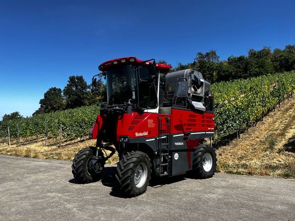 Ein Traubenlesewagen parkt neben einem Weinberg unter klarem, blauem Himmel.