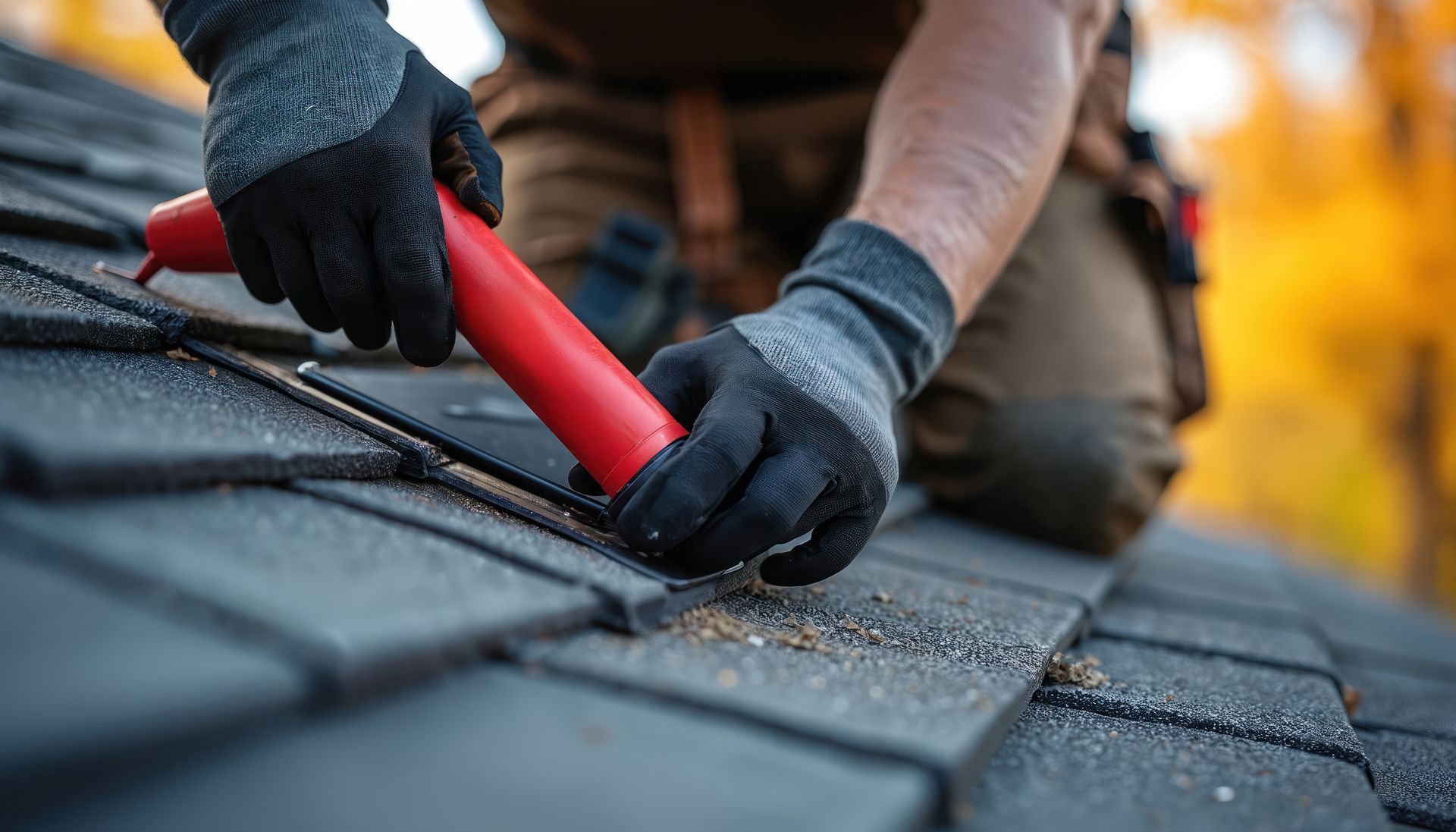 Un ouvrier portant des gants de travail utilise un outil rouge pour appliquer du mastic sur un toit en bardeaux.