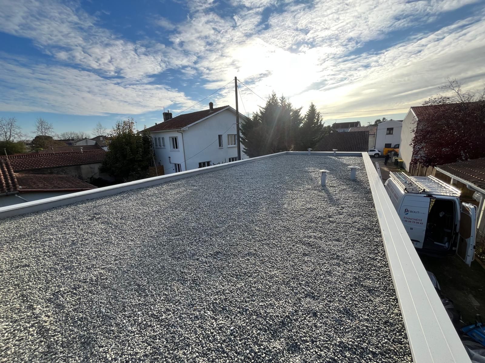 Un pinceau applique un mastic liquide foncé sur une surface plane adjacente à un rouleau de matériau de toiture gris texturé.