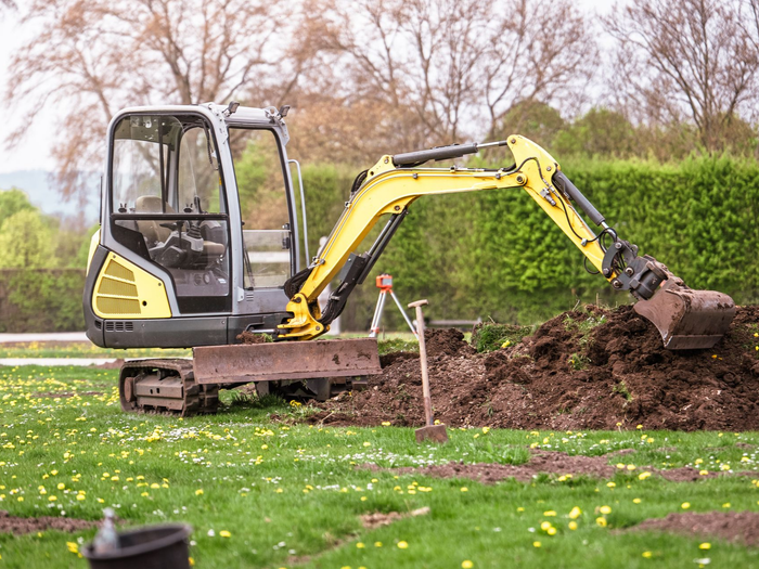 Une excavatrice jaune creuse la terre près d’une haie.