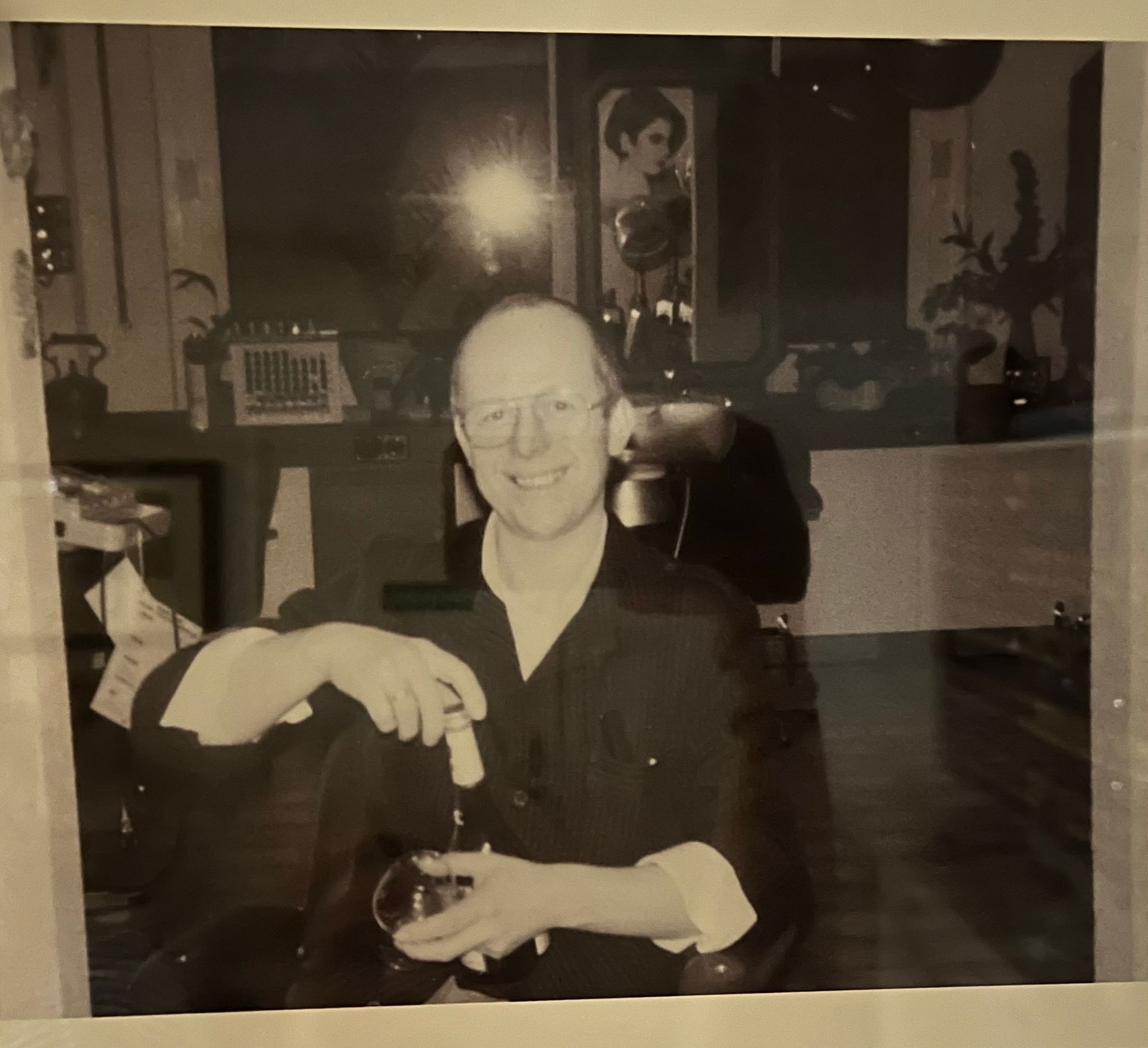 Lächelnder Mann mit Brille, Glas und Zigarre in der Hand, sitzt drinnen in einer häuslichen Umgebung.