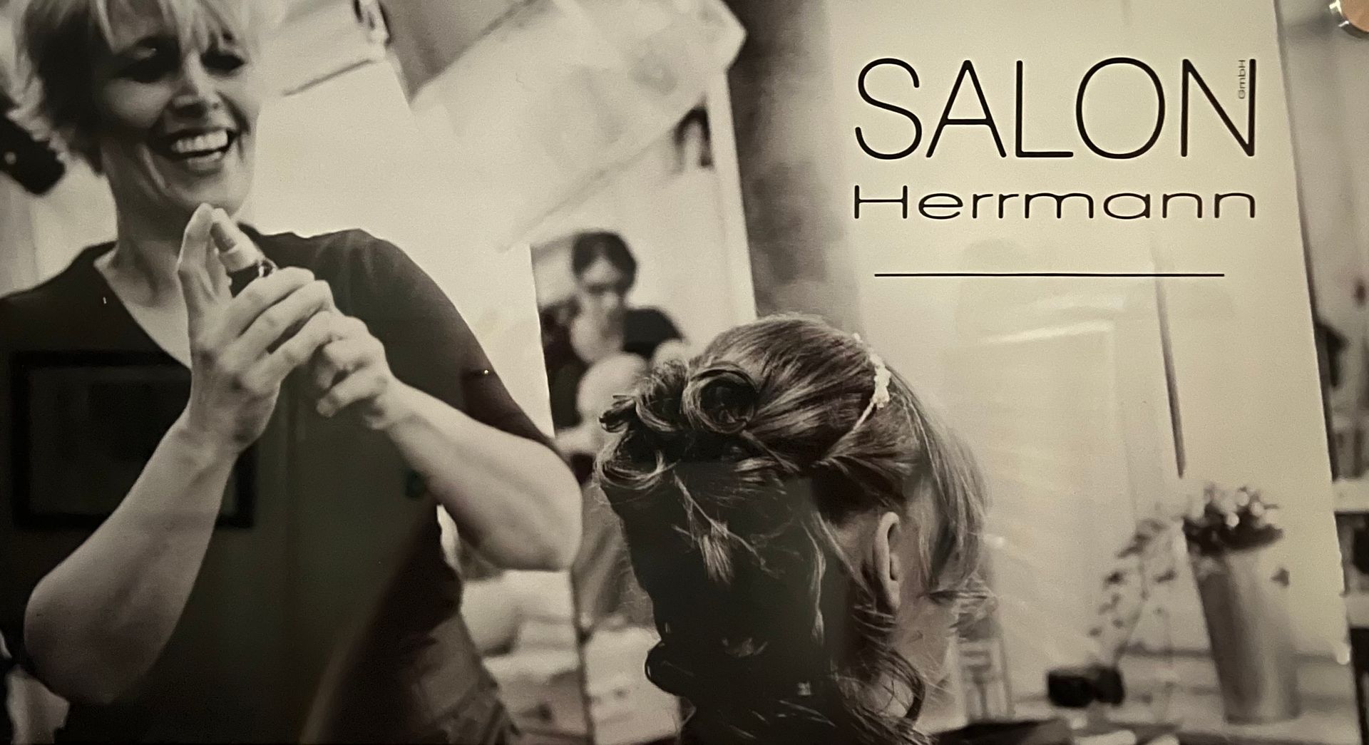 Salonmitarbeiterin besprüht lächelnd Haare; Kundin sitzt im Stuhl. Schild „SALON Herrmann“.