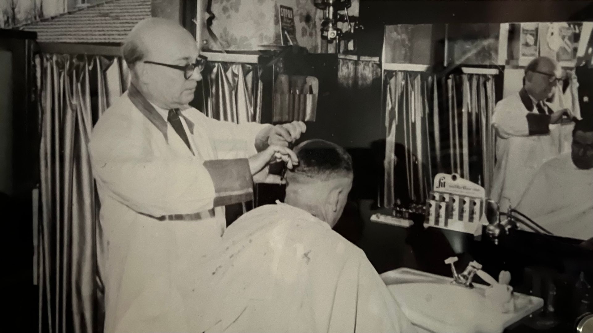 Schwarzweißfoto eines Friseurs, der einem Mann in dem Friseursalon Hermann die Haare schneidet.