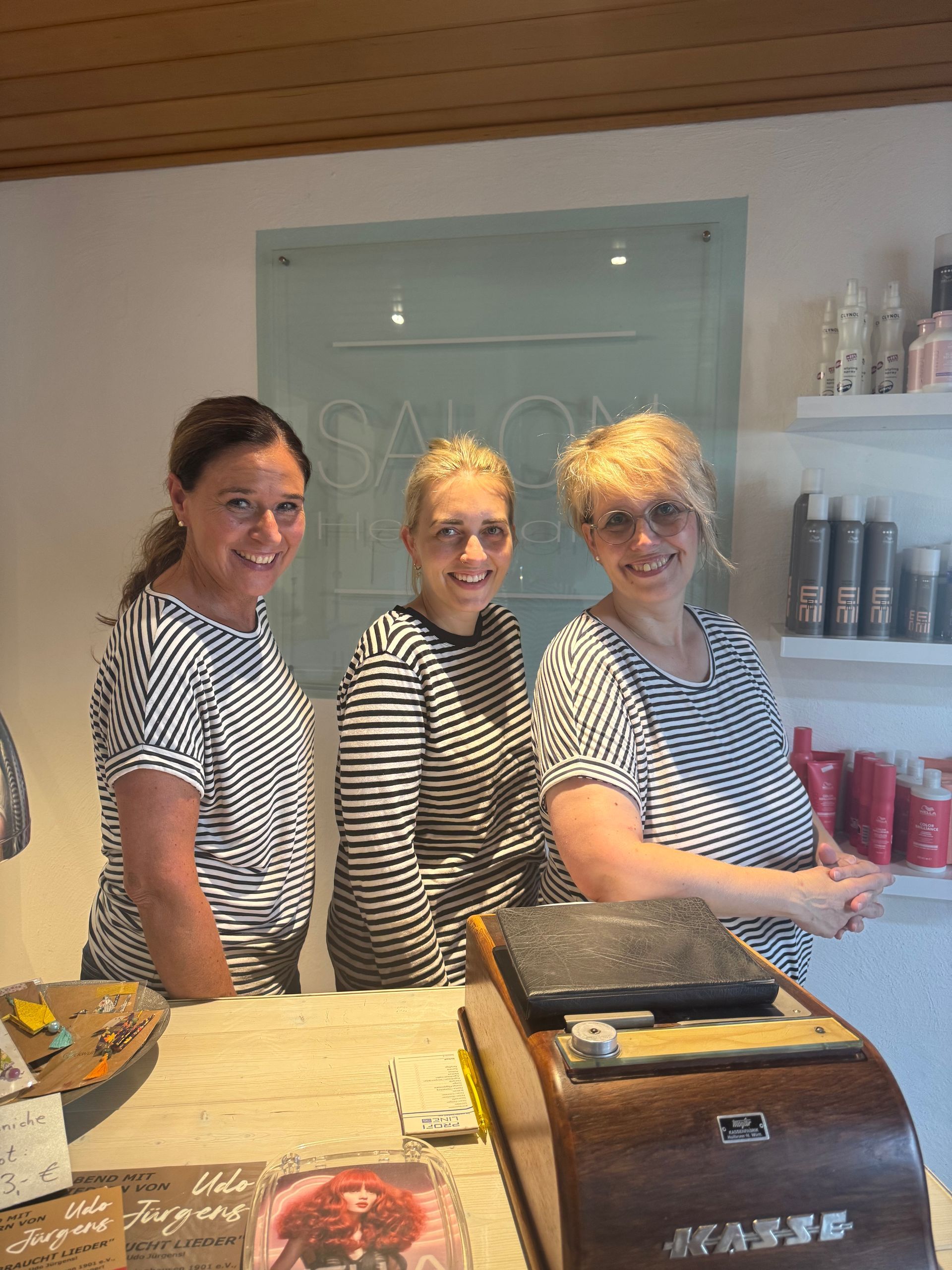 Drei lächelnde Frauen in gestreiften T-Shirts hinter einem Salontresen. Im Hintergrund ein hellblaues Salonschild.