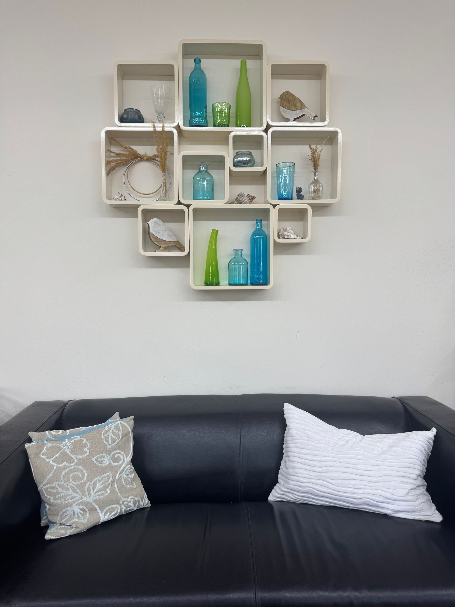 Schwarzes Sofa mit weißen und blauen Kissen unter einem an der Wand montierten quadratischen Regal mit Flaschen und Muscheln.