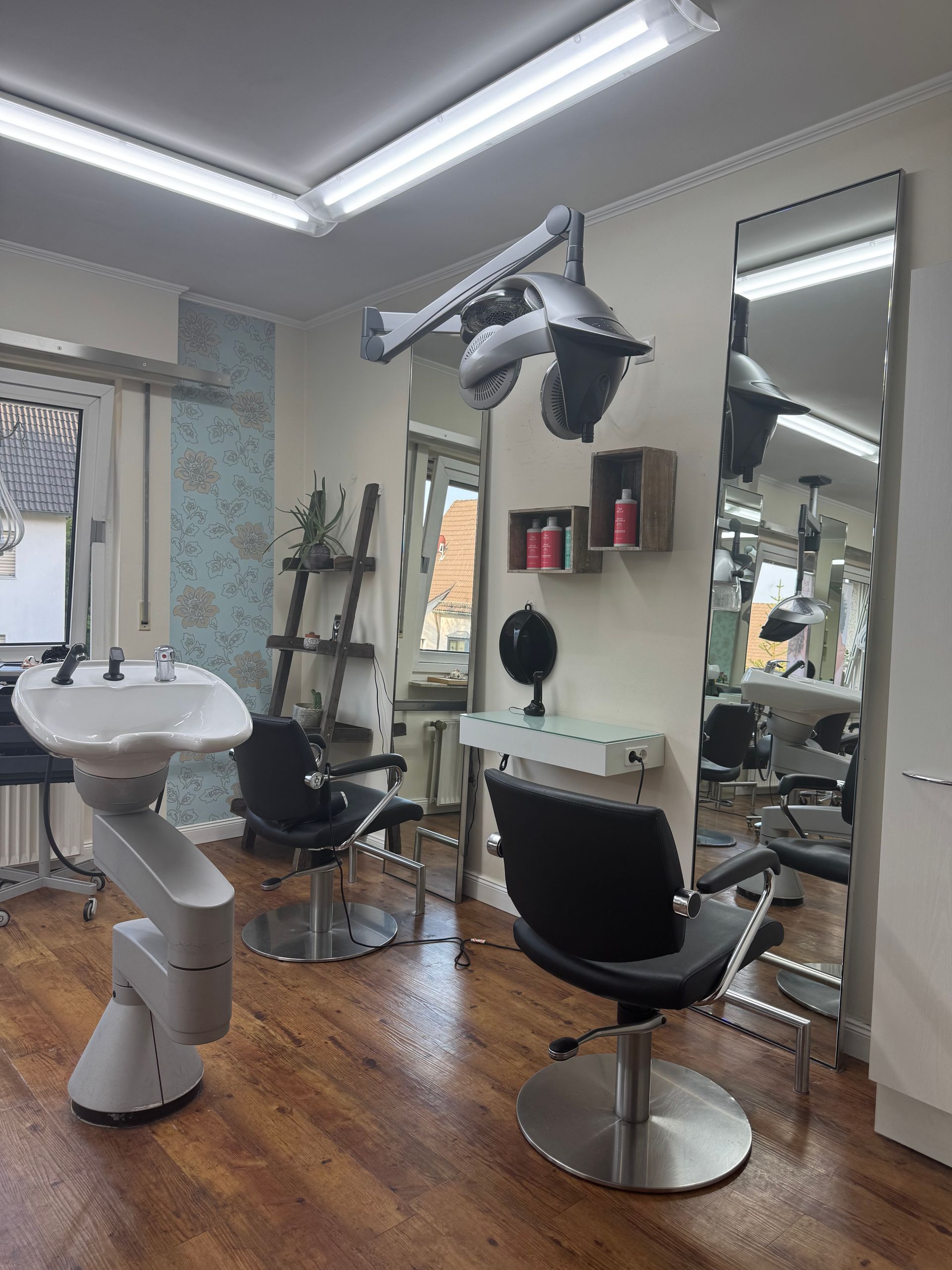 Friseursalon-Interieur mit Waschstation, Stühlen, Spiegeln und Trockner. Holzböden und helle Beleuchtung.