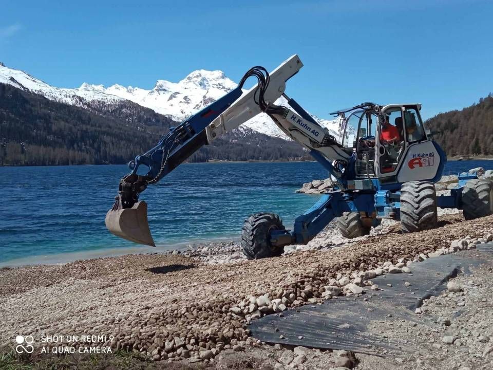 Ein blau-weisser Bagger steht am Strand neben einem See. Foto von der H. Kuhn Hoch- und Tiefbau AG