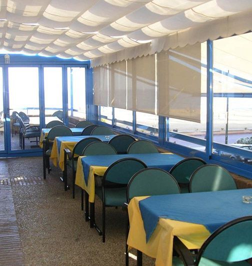 Interior de restaurante con mesas, sillas y una ventana con vistas a lo lejos. Manteles azules y amarillos.
