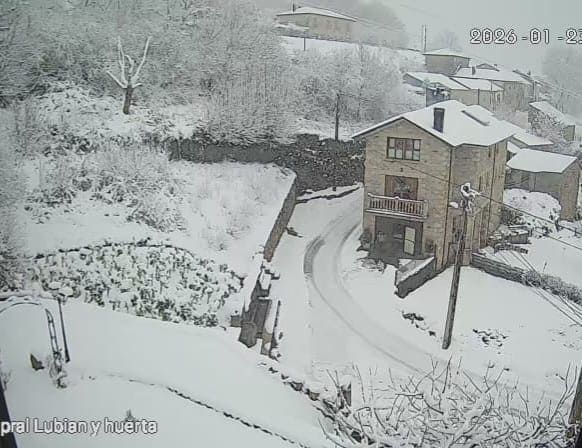 Pueblo nevado con casas de piedra y un camino sinuoso. Árboles y tierra cubiertos de blanco; nieve cayendo.