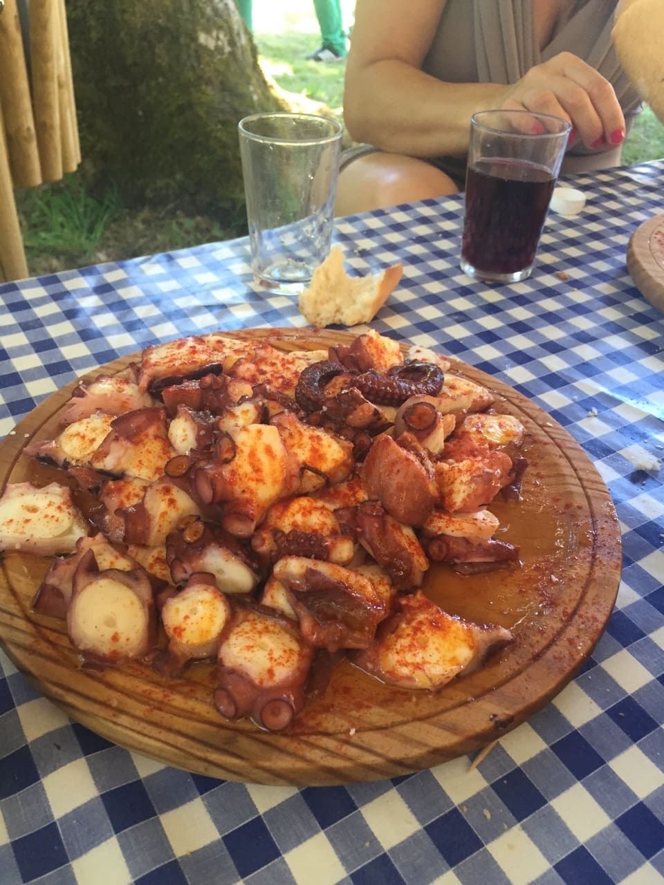 Plato de pulpo en plato de madera, espolvoreado con pimentón, sobre mantel a cuadros con bebidas.