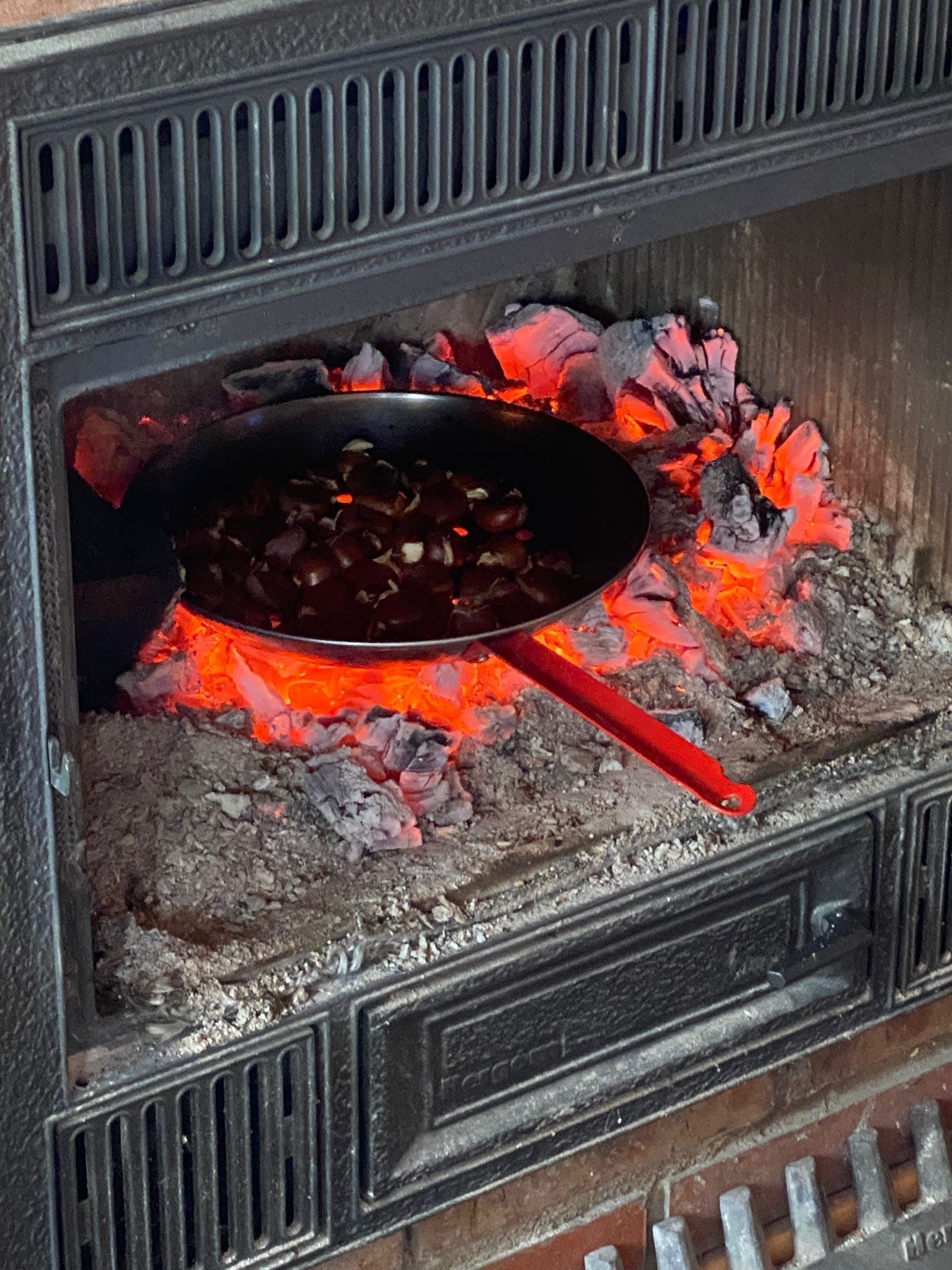 Una cacerola con comida cocinándose dentro de una chimenea sobre brasas calientes; la cacerola tiene un mango rojo.