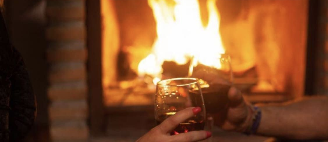 Dos personas brindan junto a una chimenea. Una mano sostiene un vaso con un líquido oscuro. Llamas al fondo.