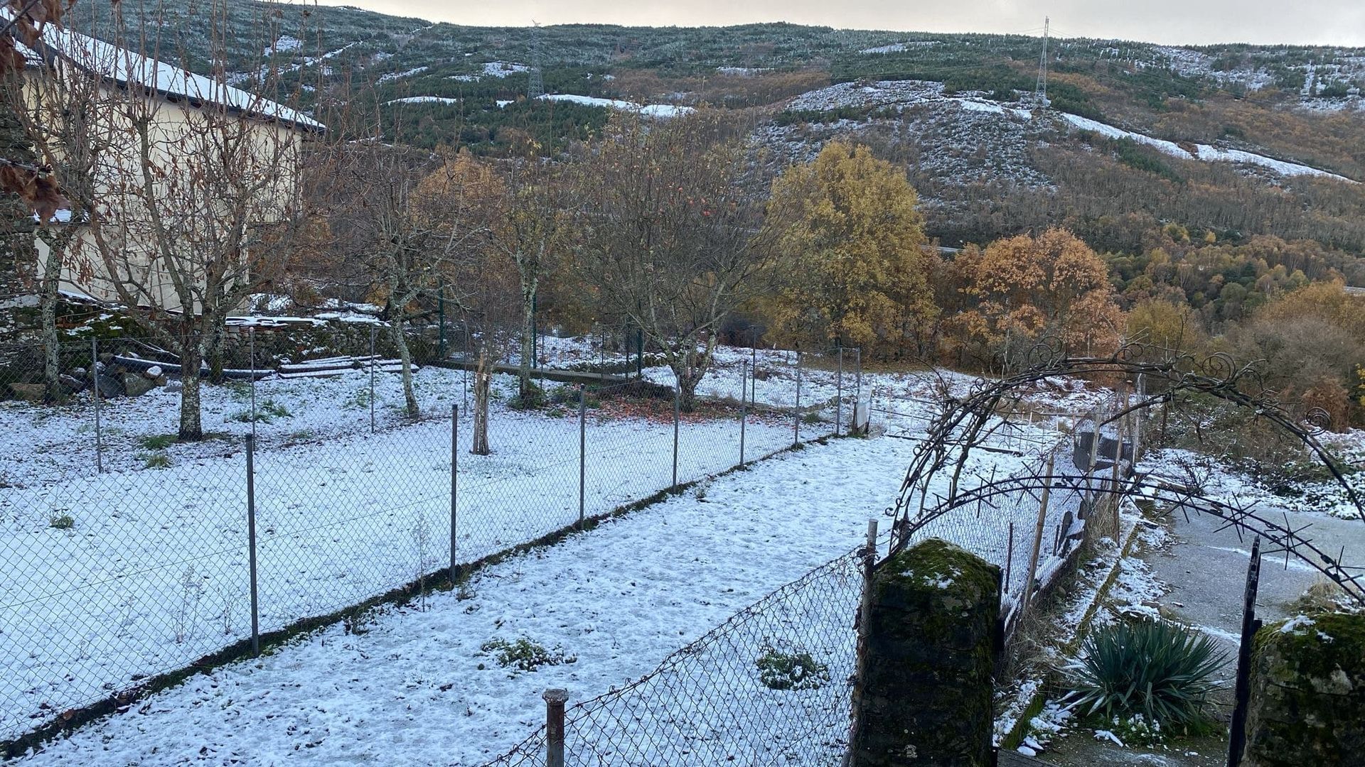 Paisaje cubierto de nieve con un edificio, árboles desnudos y una valla; colinas distantes al fondo.