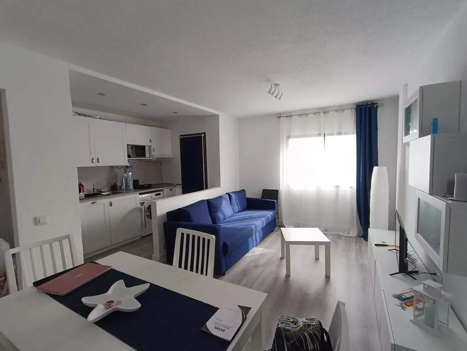 Espacio habitable de concepto abierto: cocina blanca, sofá azul, mesa de comedor y consola de TV.