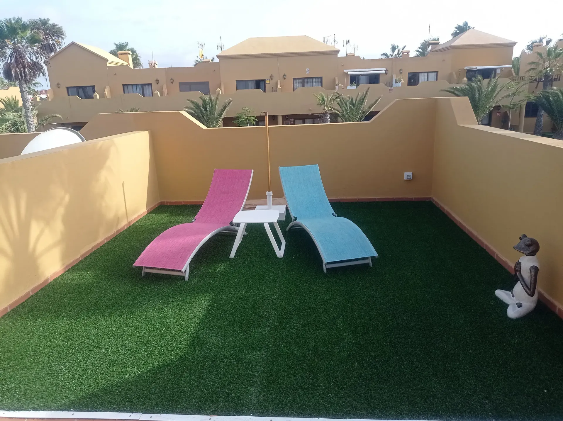 Sillones rosas y azules en un patio en la azotea con césped artificial; pequeña mesa blanca entre ellos.