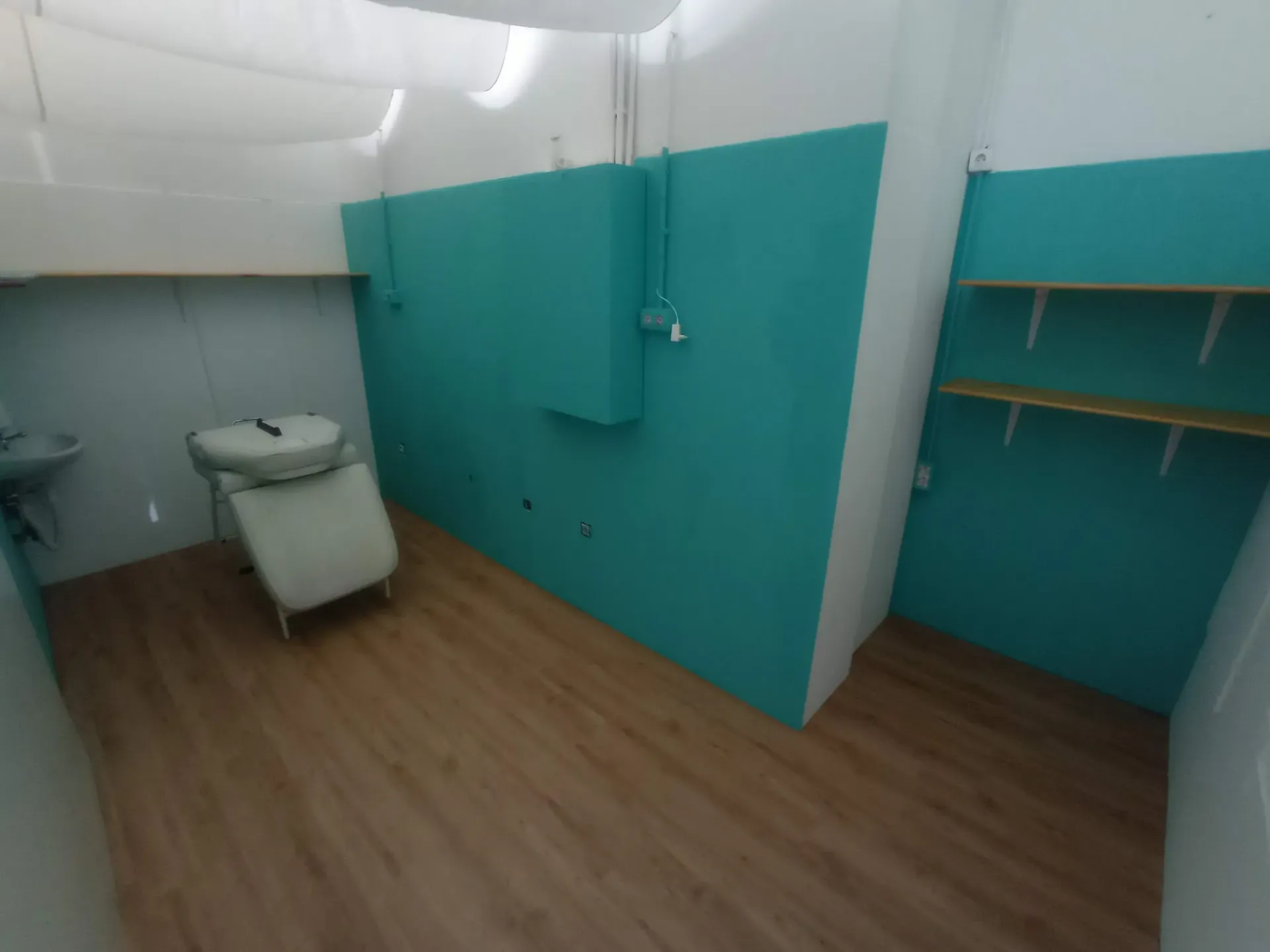 Habitación pequeña con techo blanco, pared verde azulado y suelo de madera. Incluye lavabo, silla y estanterías.