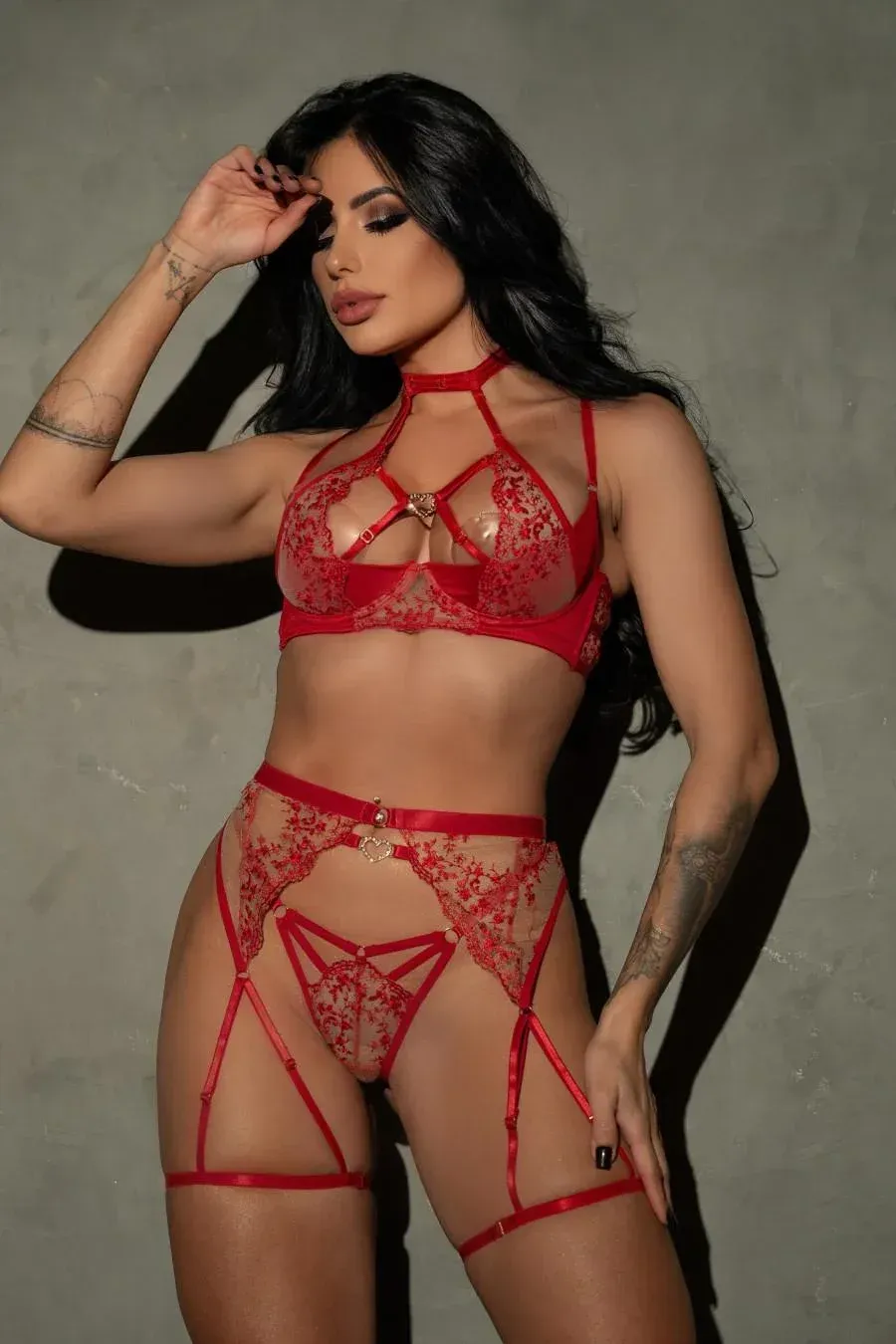 Mujer en lencería roja, con las manos en la cabeza, de pie cerca de una pared gris.