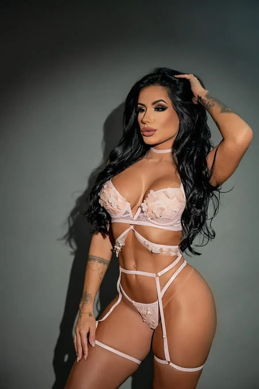 Mujer en lencería rosa pálido, con la mano en el cabello, posando sobre un fondo gris.