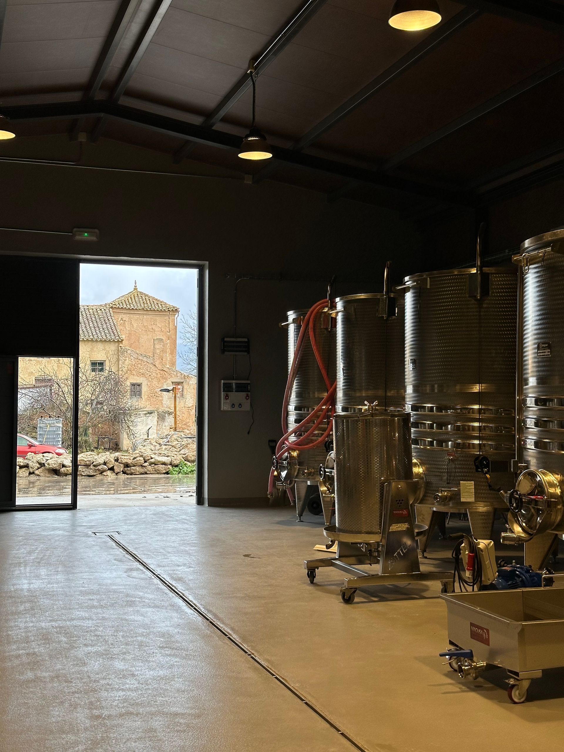 Bodega con grandes depósitos de acero inoxidable y una puerta abierta que deja ver un edificio de piedra en el exterior.