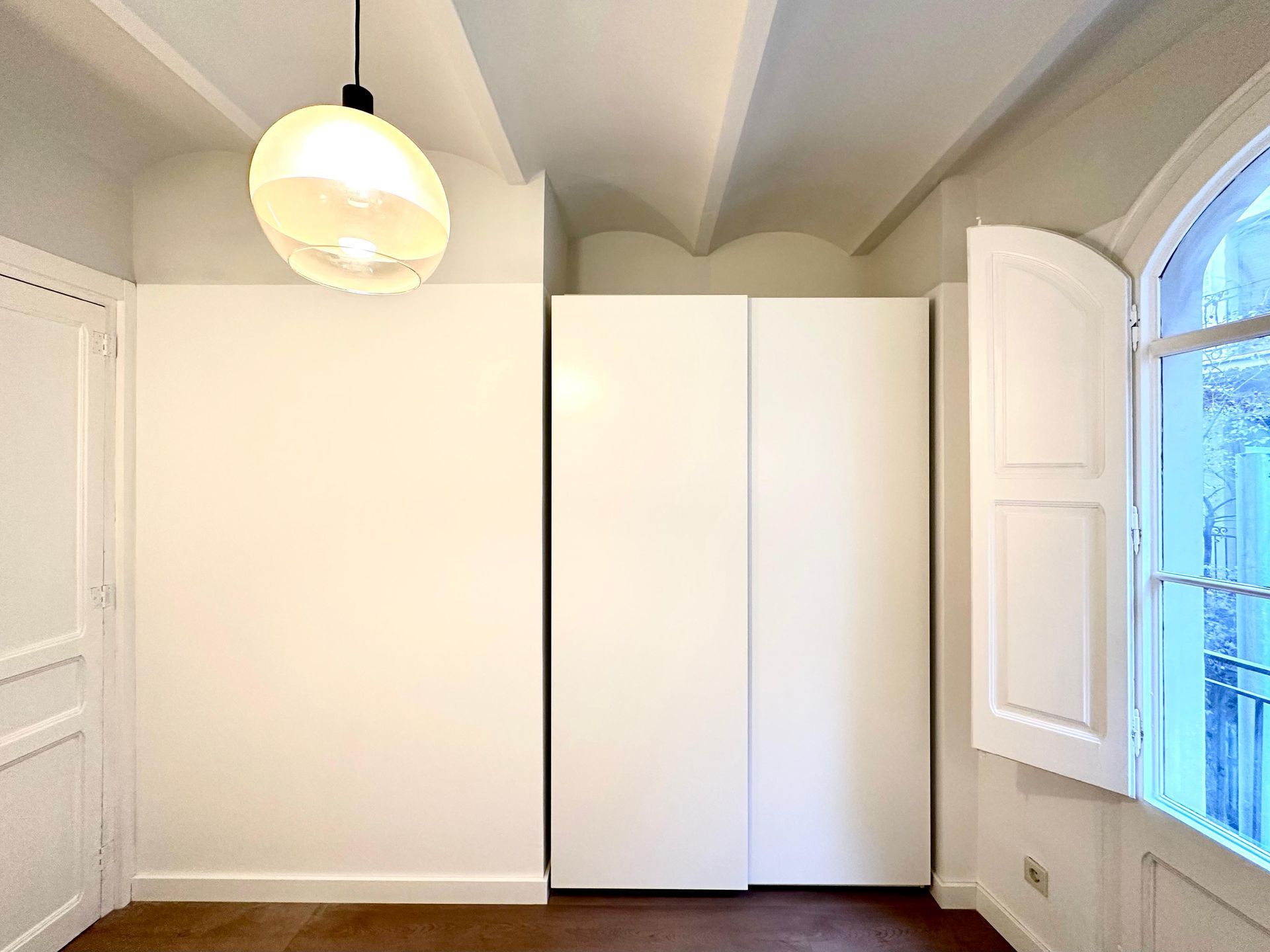 Habitación luminosa y vacía con paredes blancas, armario empotrado, ventana arqueada y lámpara colgante.