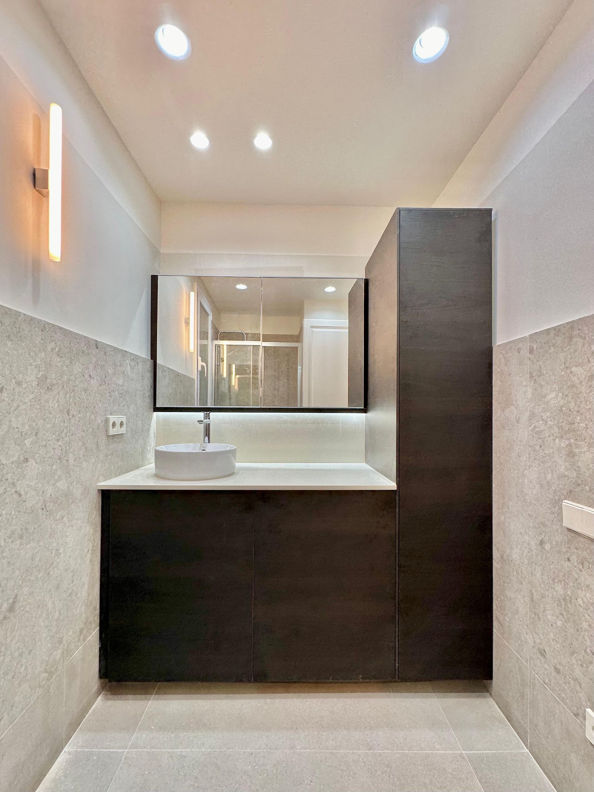 Tocador de baño moderno con espejo, lavabo, apliques de pared y armarios de madera oscura.