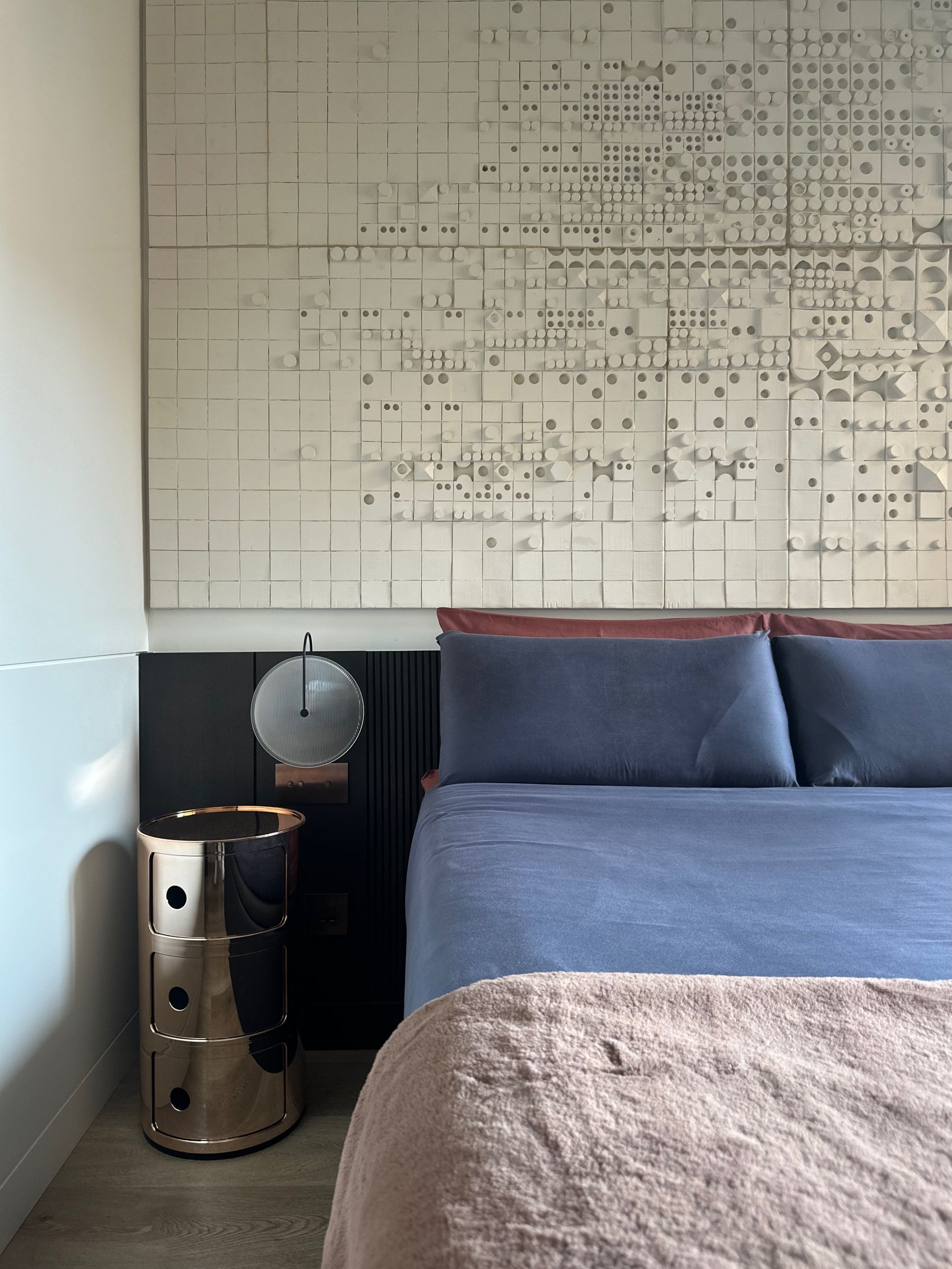 Cama azul junto a una mesita auxiliar plateada y una lámpara debajo de un gran panel de arte abstracto en la pared.