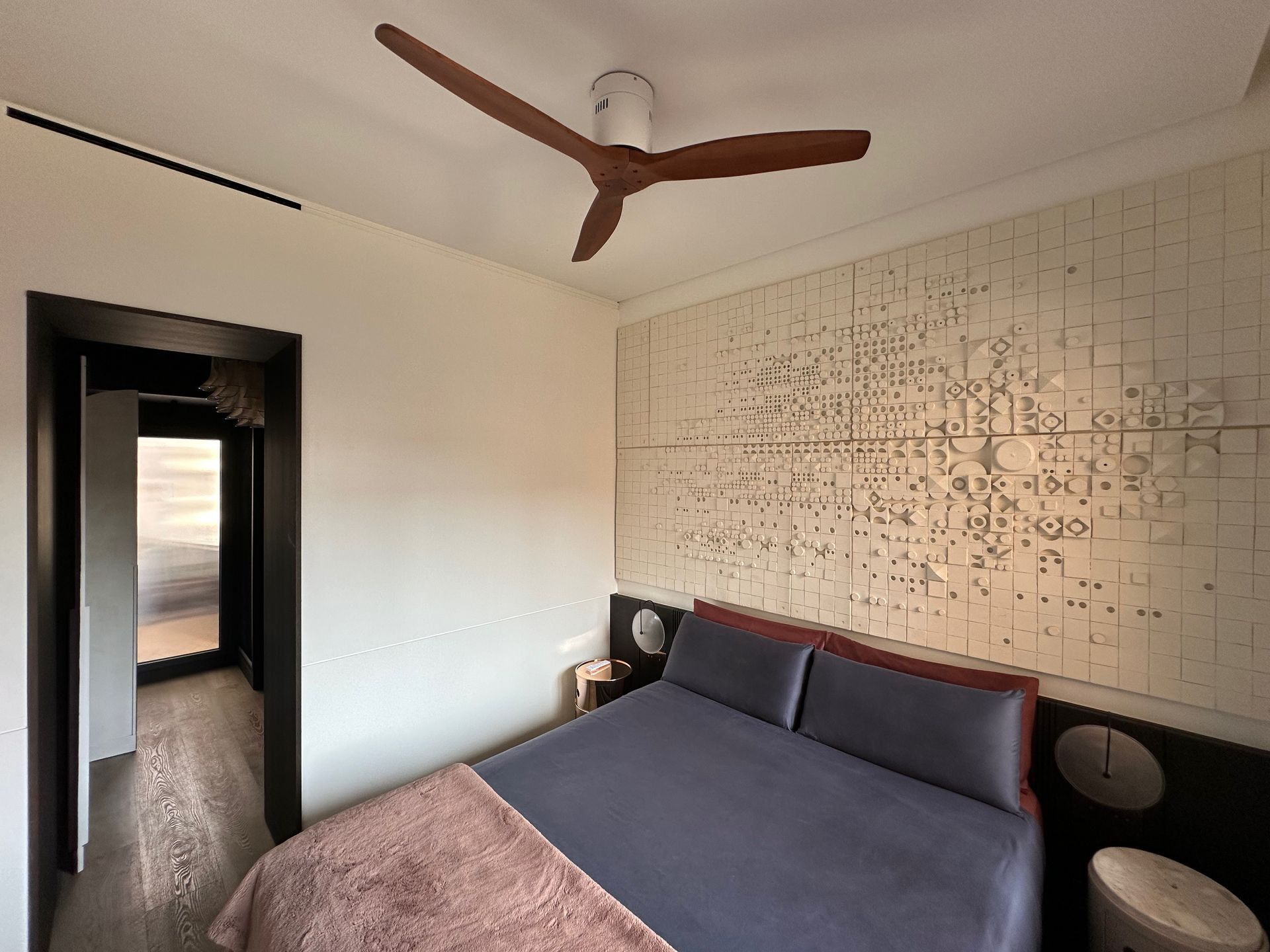 Dormitorio con una cama grande, ventilador de techo y una pared decorativa con estampado, visto desde la puerta.