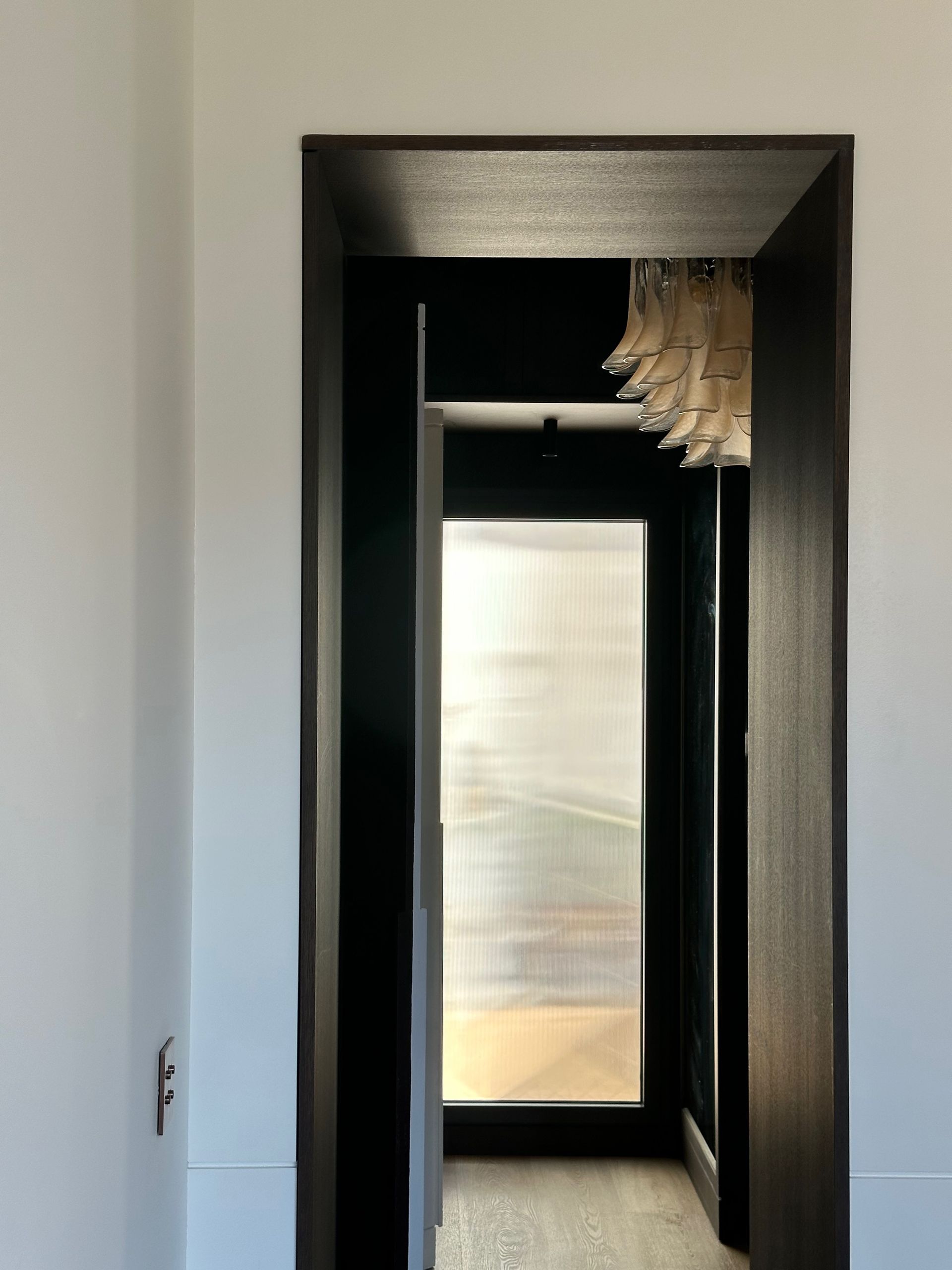 Pasillo estrecho con una puerta oscura que enmarca una ventana luminosa al final.