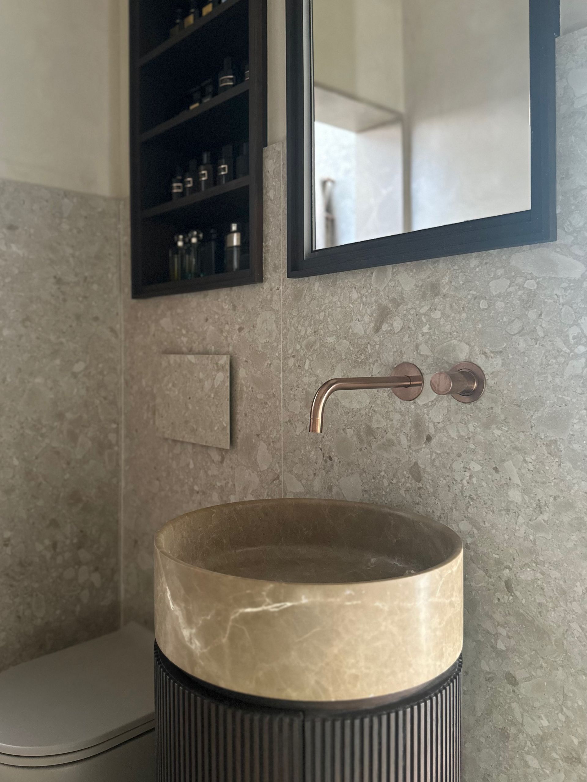 Baño moderno con lavabo de piedra, grifo de cobre, espejo y estante de pared para botellas.