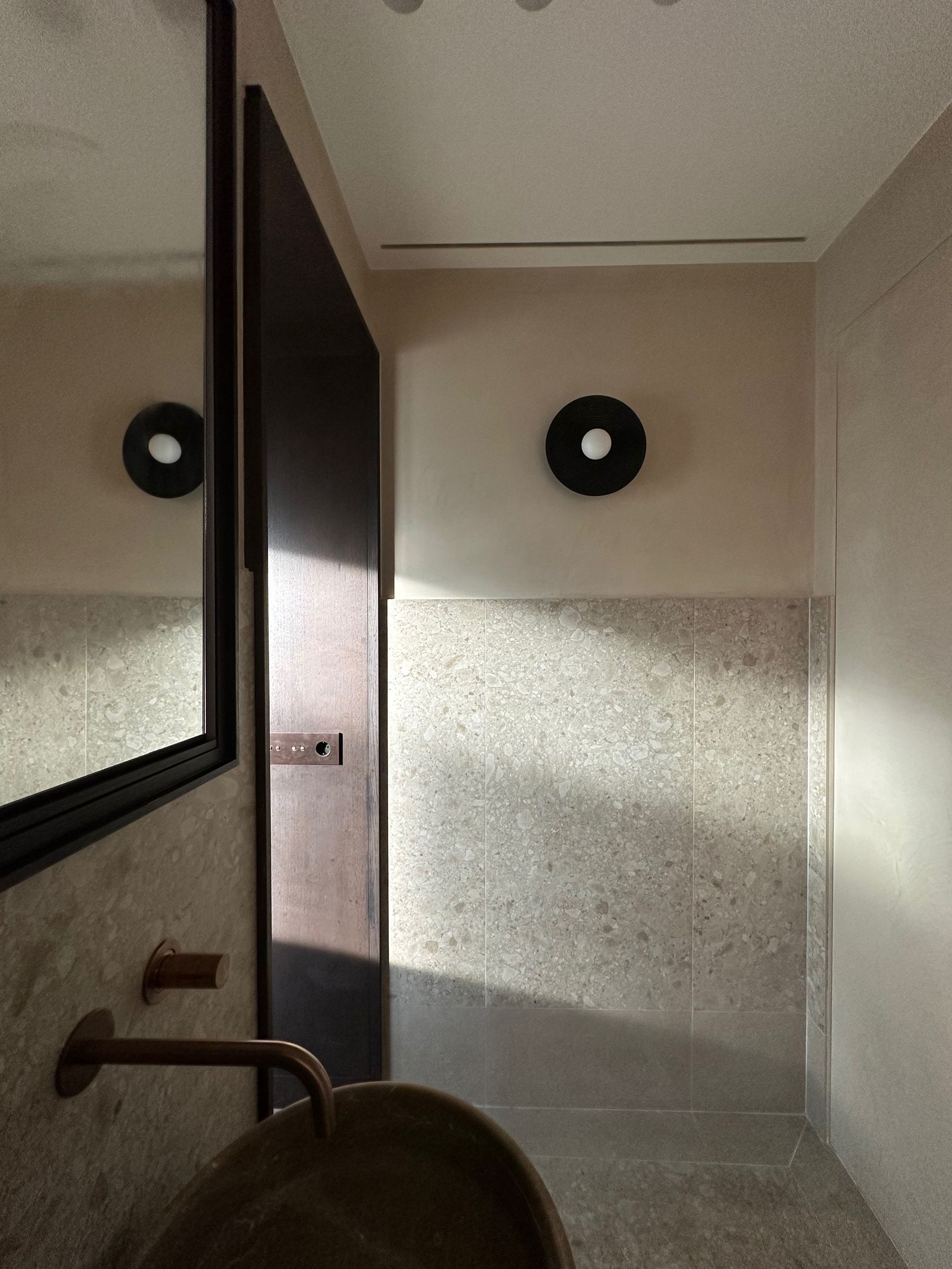 Baño pequeño con paredes beige, espejo, grifo en el lavabo y una puerta oscura abierta.