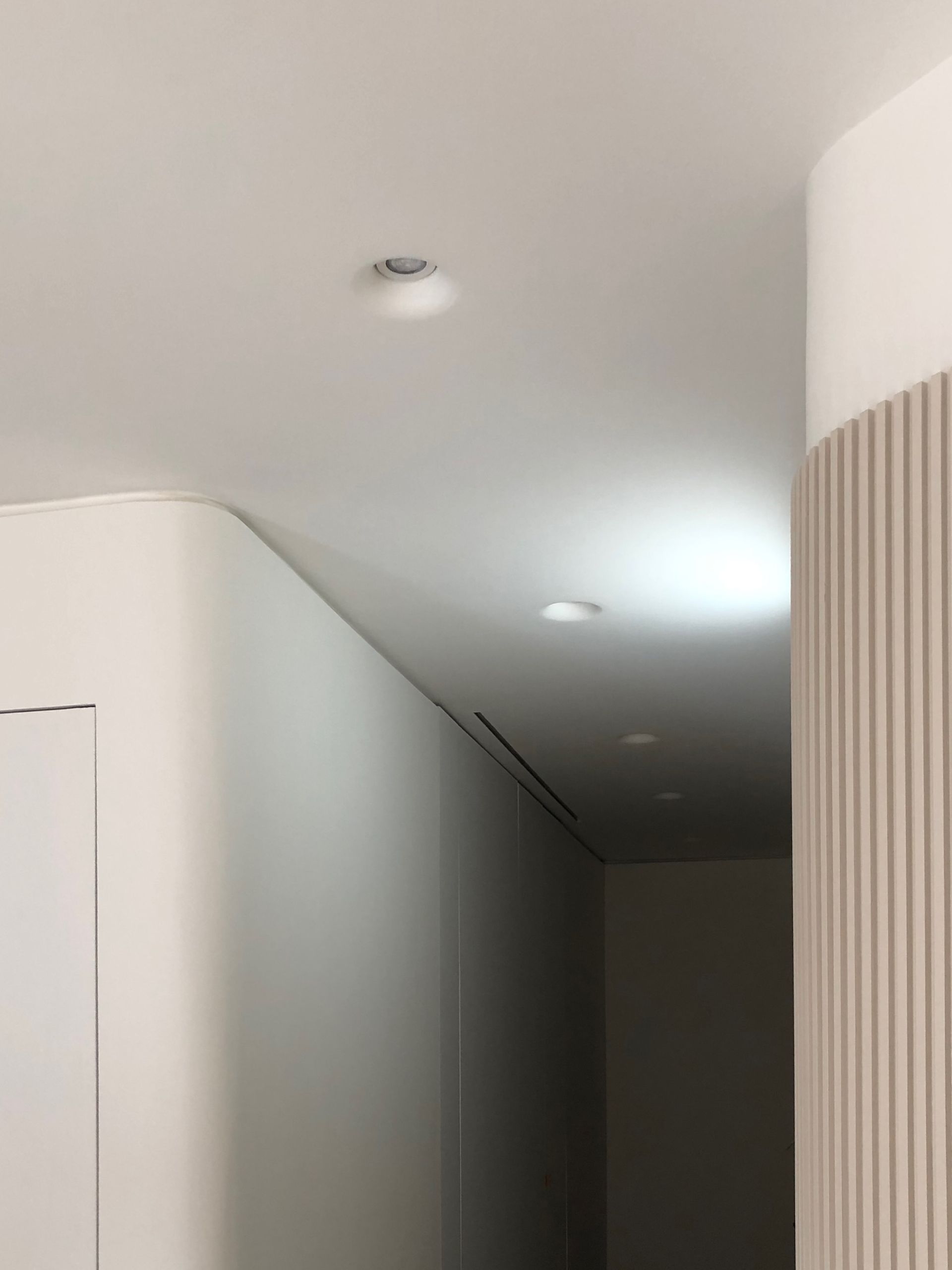 Pasillo moderno con paredes blancas, luces empotradas en el techo y un panel decorativo curvo de listones de madera.