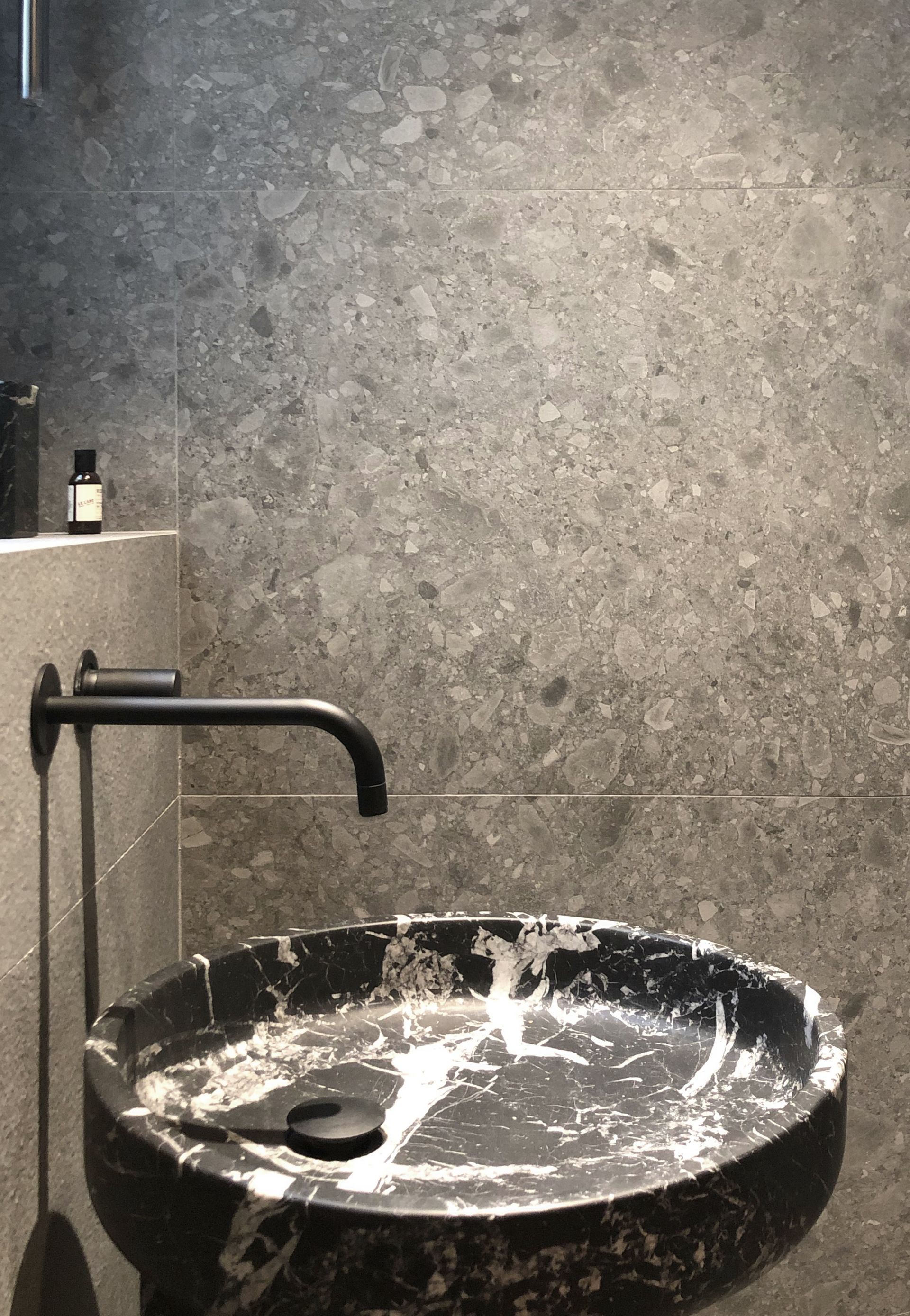 Lavabo negro con grifo sobre una pared de baño alicatada en gris, con salpicaduras de agua en el interior.
