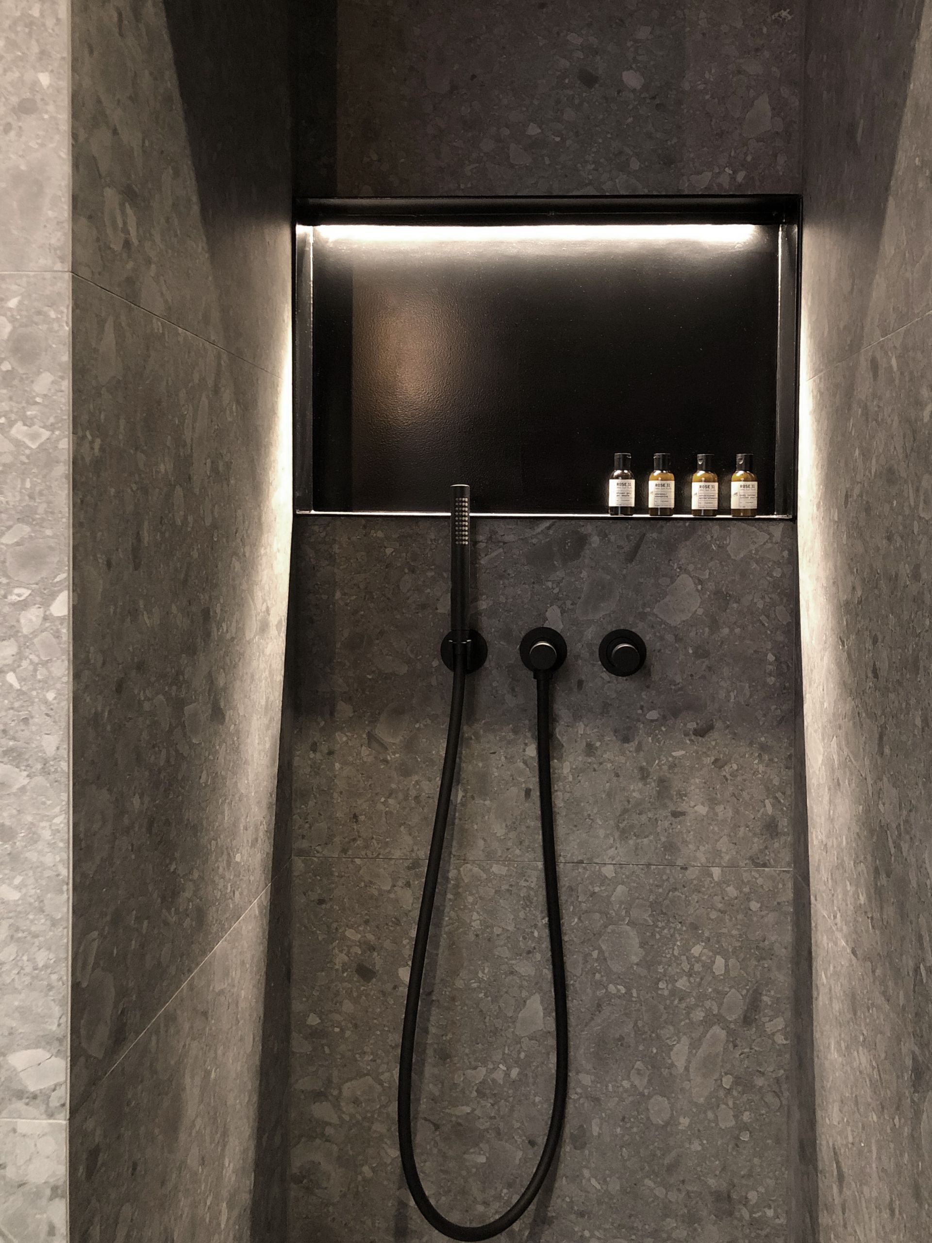 Ducha minimalista con paredes de piedra oscura, grifería negra y botellas pequeñas en una repisa.