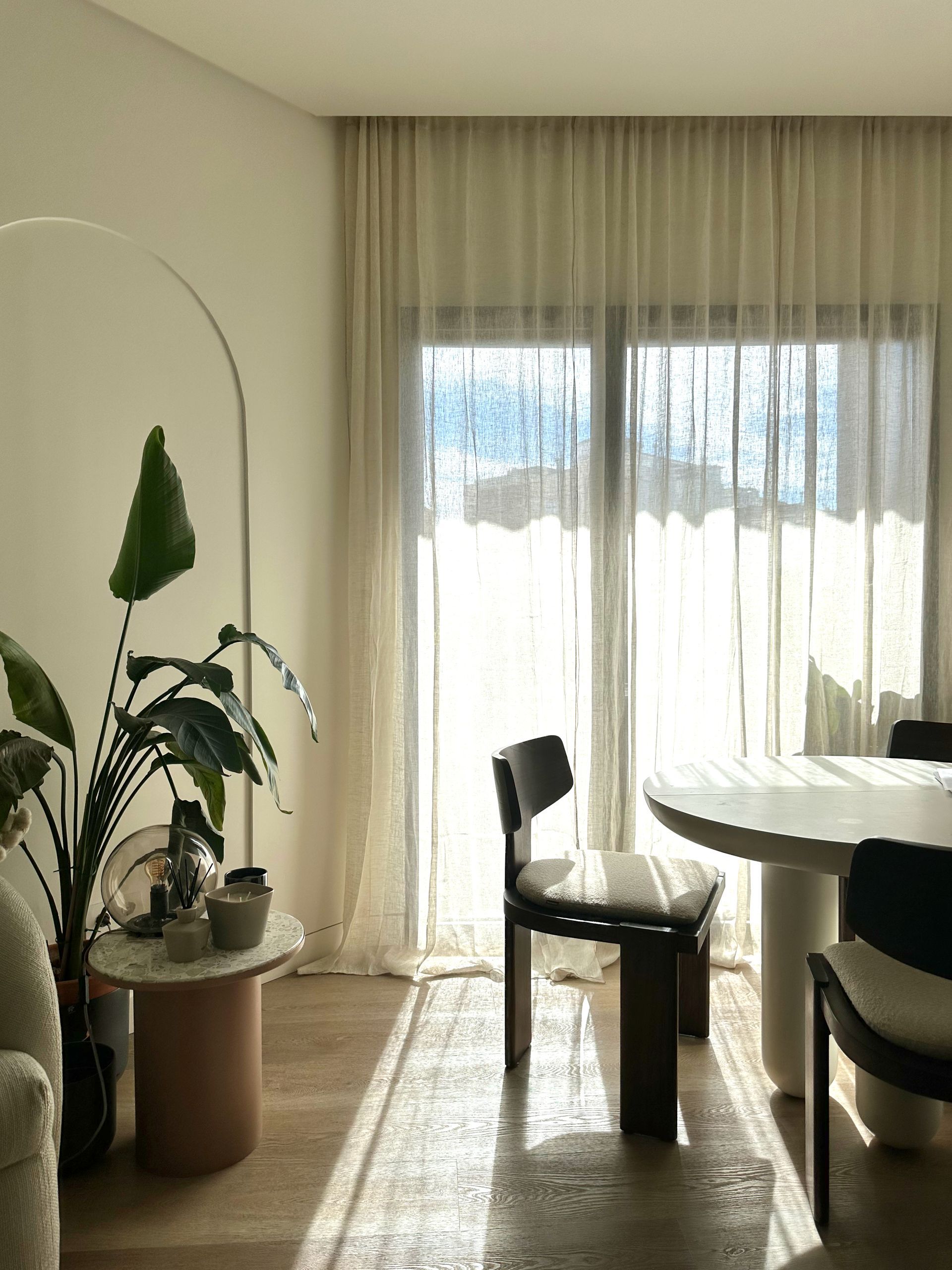 Comedor luminoso con cortinas transparentes, silla negra, mesa redonda y plantas en macetas.