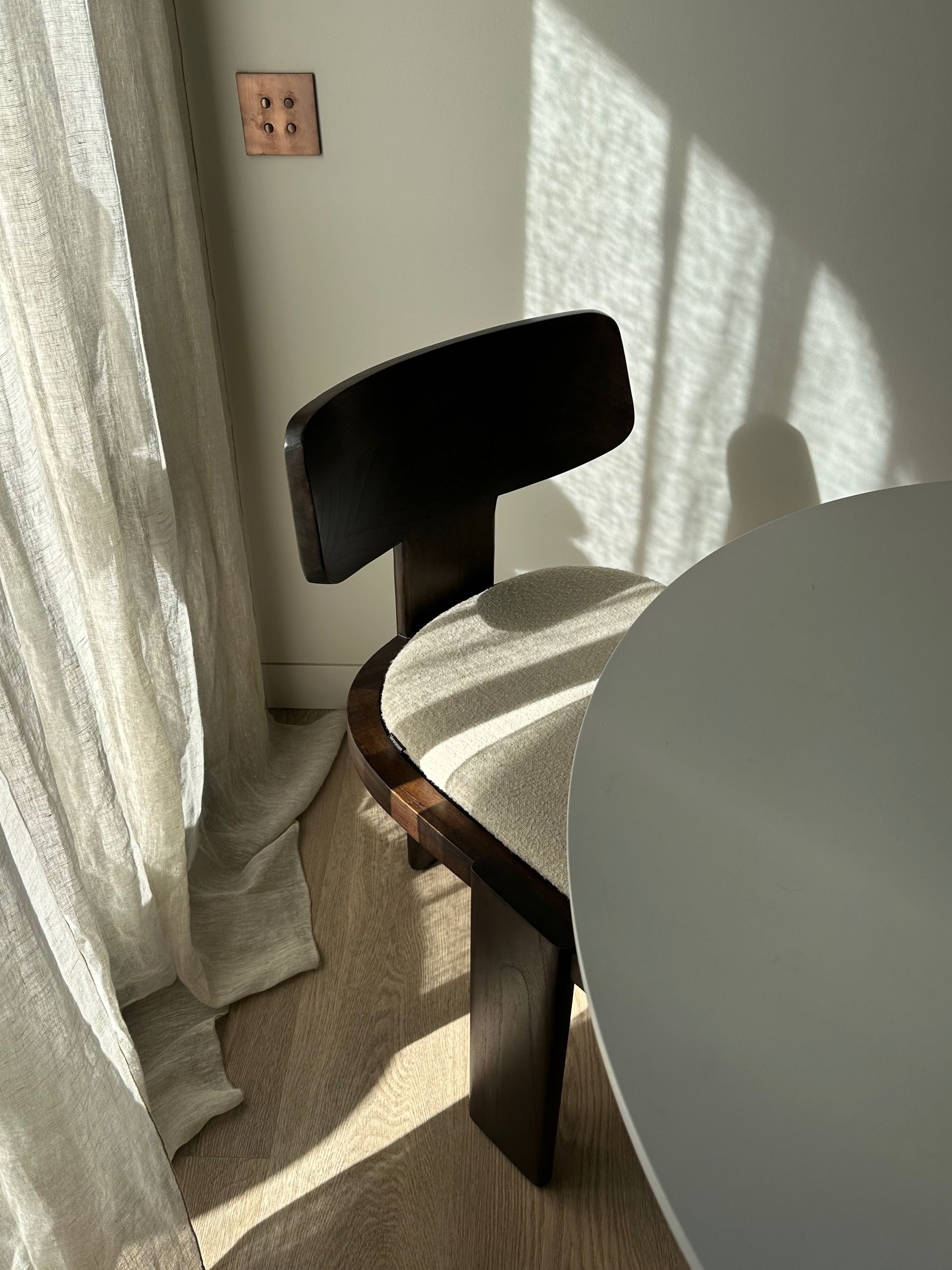 Silla de comedor iluminada por el sol junto a una mesa redonda y una ventana con cortinas en una habitación luminosa.