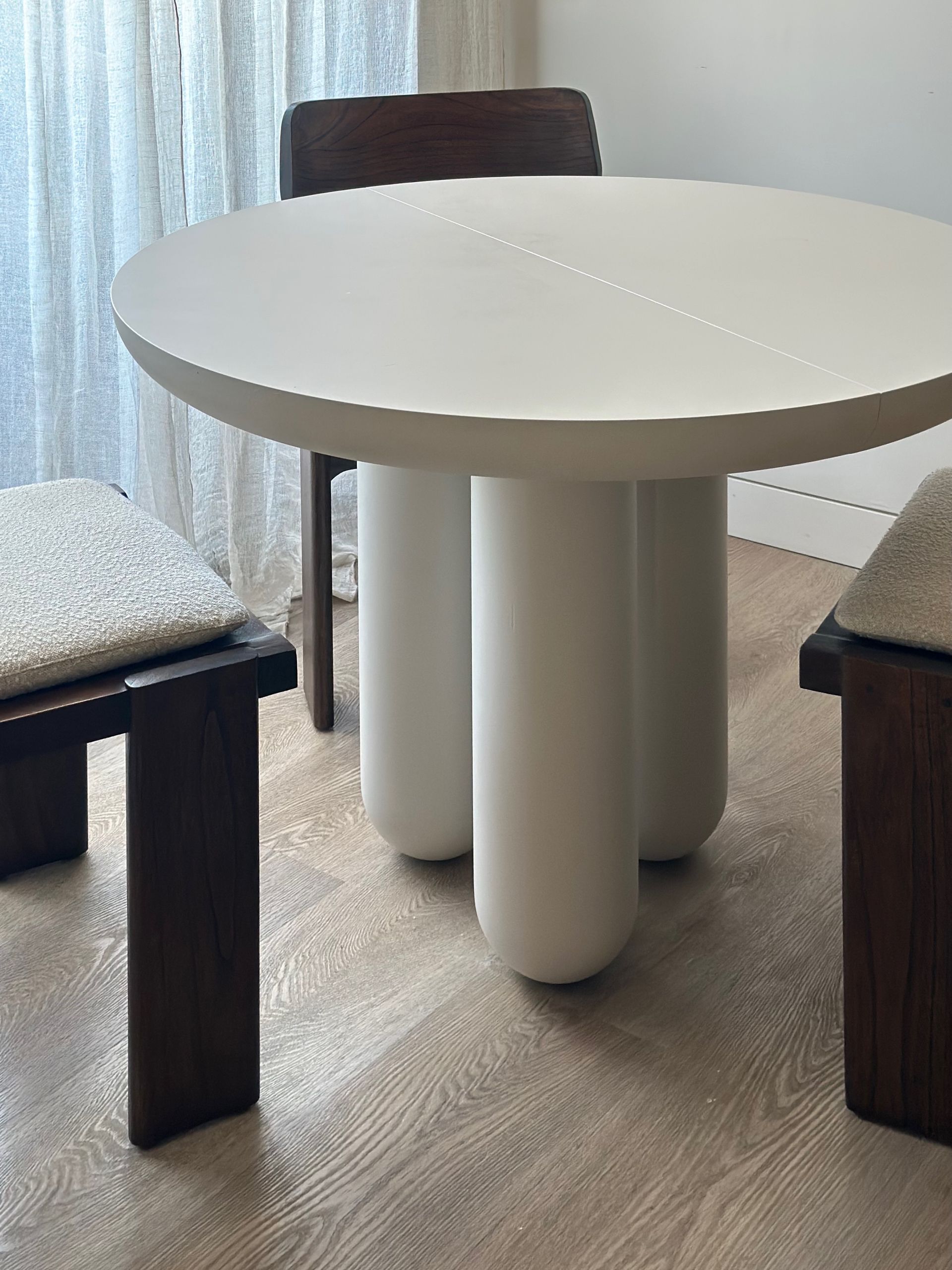 Mesa redonda blanca con base cilíndrica entre sillas de madera oscura en una habitación luminosa.