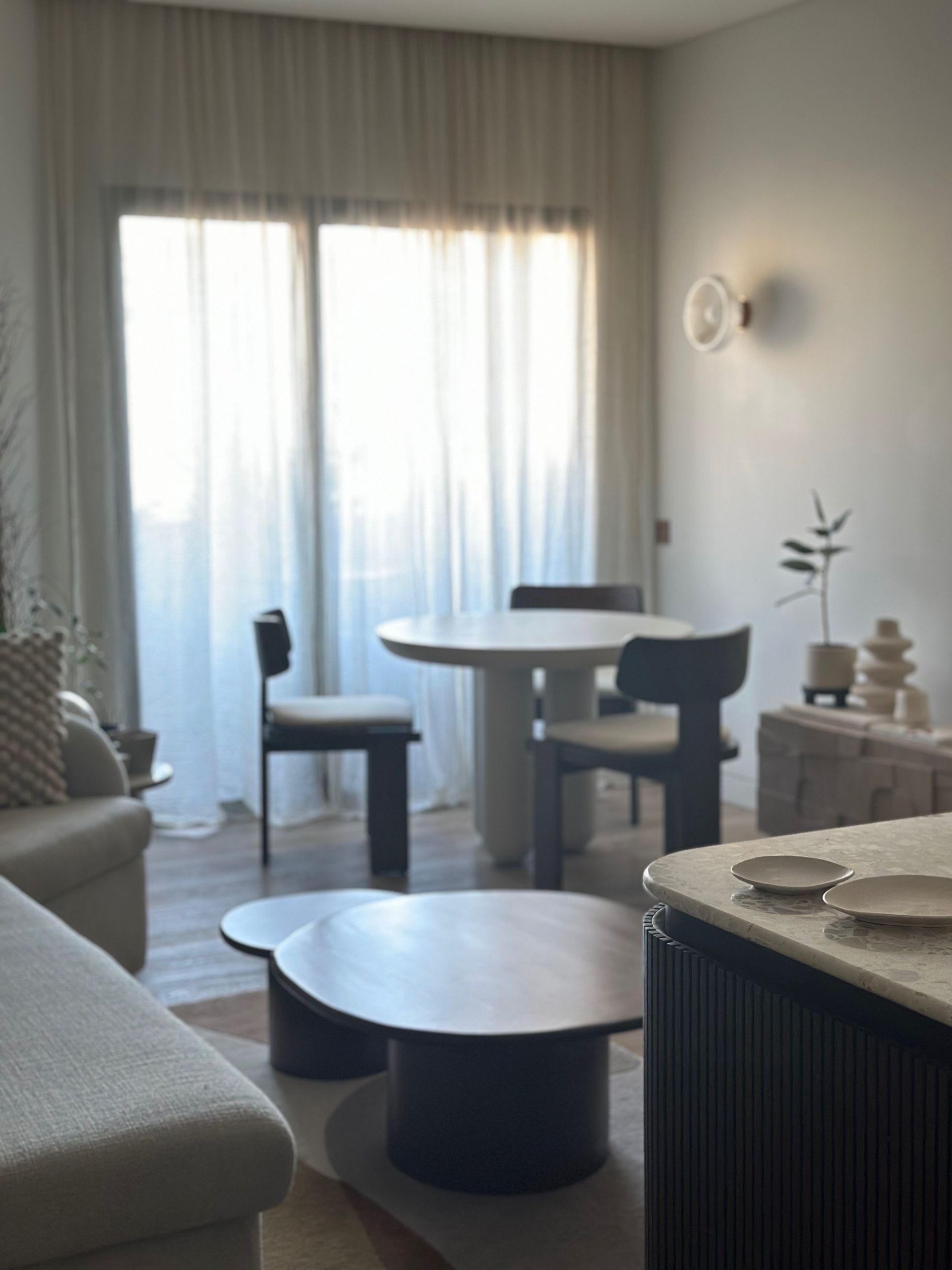Sala de estar minimalista con mesa redonda, dos sillas, sofá y cortinas transparentes junto a una ventana luminosa.