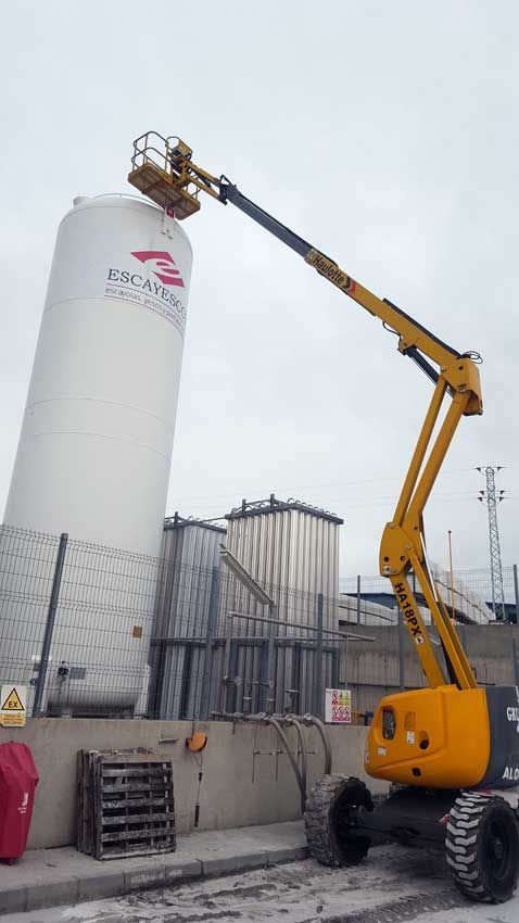 Un elevador aéreo amarillo está sentado frente a un gran tanque blanco.