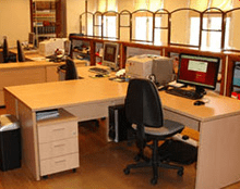 Cubículo de oficina con escritorio, silla, monitores de computadora y ventanas al fondo.