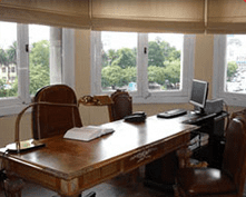 Oficina en casa con escritorio de madera, silla, computadora portátil, papeles y grandes ventanales con vista a zonas verdes.