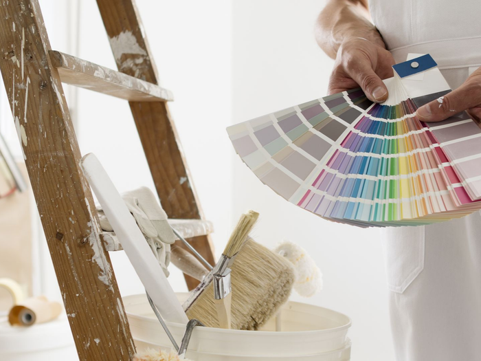 Une personne tenant un échantillon de couleur se trouve à côté d'une échelle et d'un seau de peinture dans une pièce.