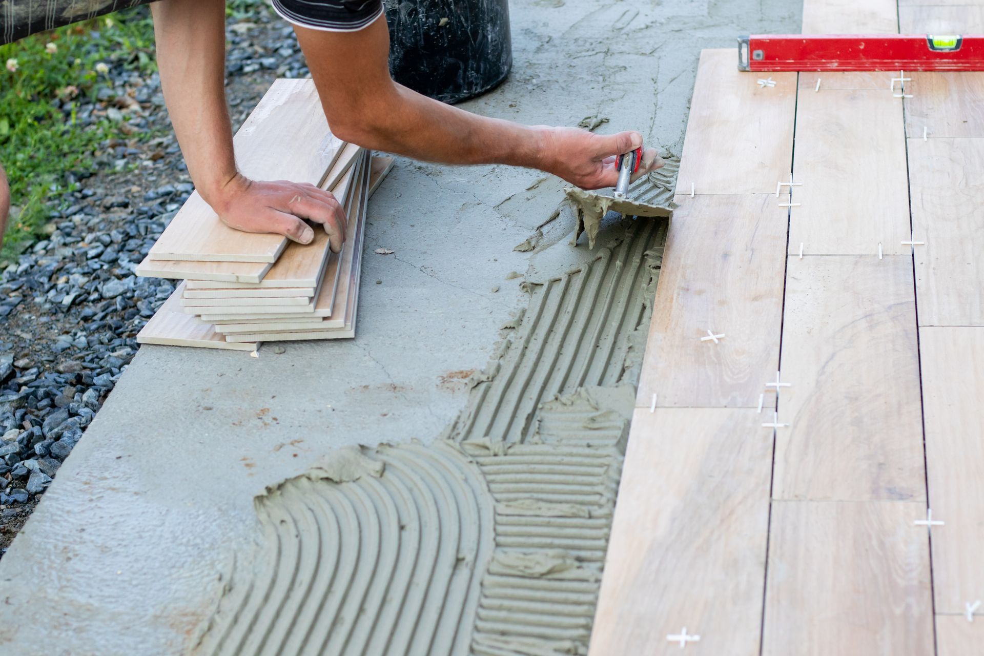 Des mains appliquent de la colle à carrelage grise à côté de carreaux de sol beiges déjà posés à l'extérieur.