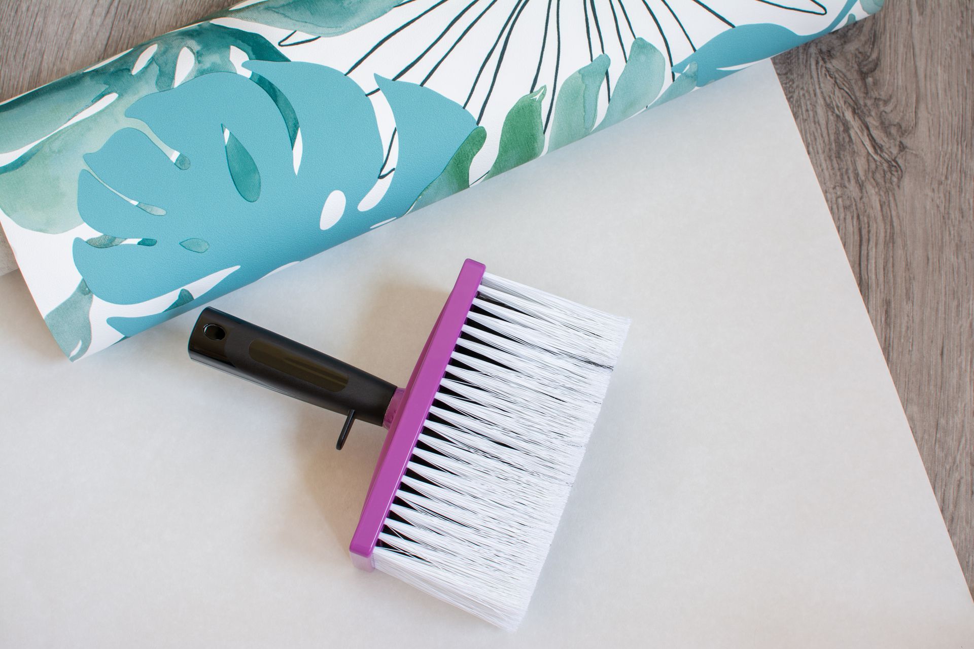 Pinceau blanc et violet sur une surface blanche, à côté d'un rouleau de papier peint à motifs floraux.